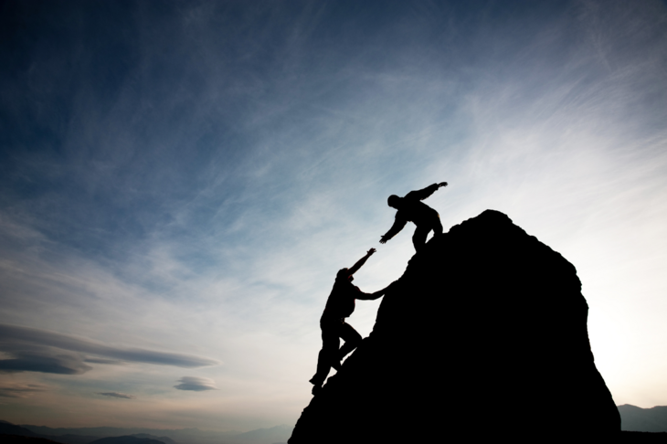 To mennesker på toppen av et fjell. Det ene menneske strekker ut armen for å hjelpe den andre opp.