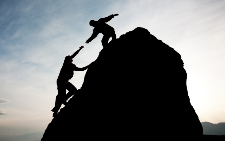 To mennesker på en fjelltopp i solnedgangen. Den ene personen rekker ut hånden for å hjelpe den andre opp til toppen.